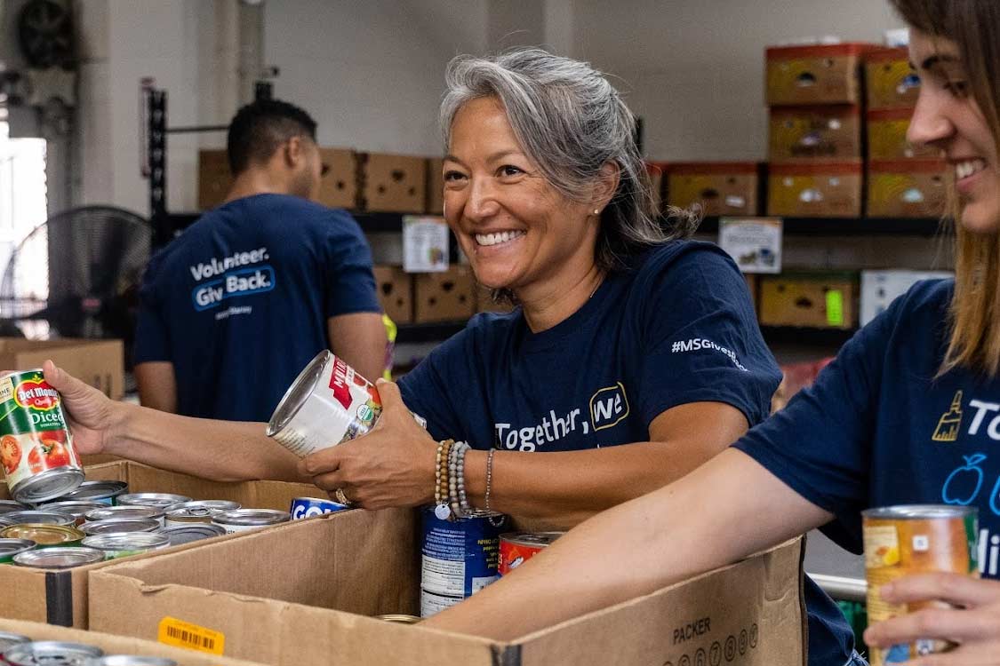 A cheerful volunteer is sorting canned goods in a warehouse, with other volunteers working in the background, all wearing blue shirts with the text "Together, We Can."
