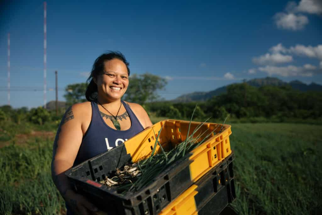 A cheerful hawaiian woman holding a crate of green onions in a sunny field with mountains in the background.